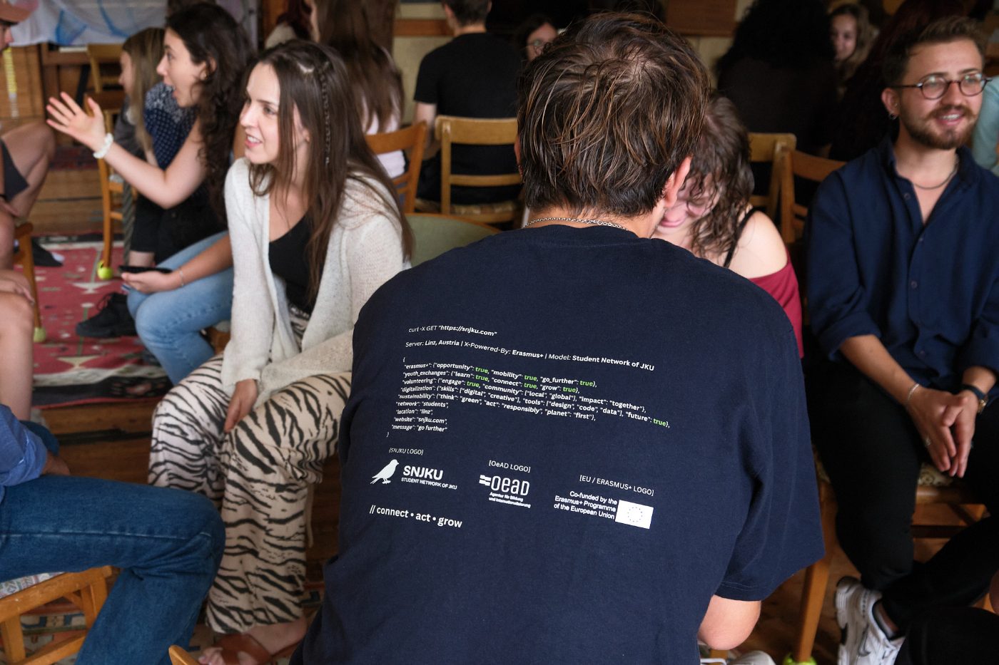 Participants sitting in a circle, one wearing an SNJKU shirt with the project logos